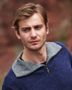 A young man with light brown hair wearing a blue sweater outdoors.