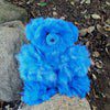A vibrant blue teddy bear sitting outdoors on some rocks.